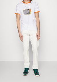Hvid t-shirt med orange kantet krave, prydet med et retro tv-grafik, parret med off-white bukser og tealfarvede sneakers.