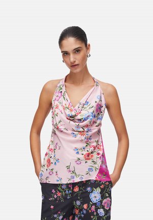 Donna che indossa una canotta rosa fantasia floreale senza maniche e pantaloni neri con motivo floreale, in piedi con le mani nelle tasche davanti a uno sfondo semplice.