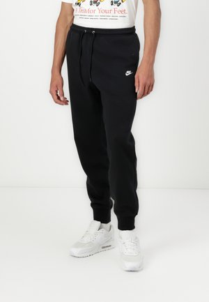 Persoon die zwarte Nike joggingbroek met trekkoord en witte sneakers draagt, staand tegen een effen witte achtergrond.