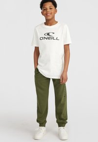 Camiseta blanca de algodón con el logo negro de "O'Neill", combinada con joggers de color verde oliva. Zapatillas blancas completan el conjunto. Diseño simple y casual.