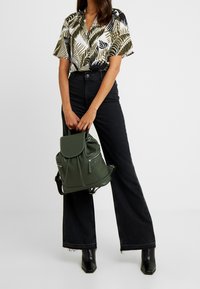 Sac à dos en cuir vert avec un rabat et une fermeture à cordon, comportant plusieurs poches zippées. Porté avec un jean évasé taille haute noir.