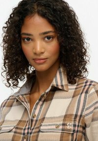 Camisa de cuadros marrón con dise�ño abotonado, con detalles en beige y naranja, cuello gris y un pequeño logo blanco en el bolsillo.