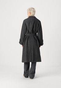 Abrigo largo gris de espiga con cinturón de lazo, grandes solapas y puños abotonados, exhibido desde la parte posterior. Lleva sobre pantalones grises de pierna ancha.