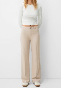 Beige vida byxor med en mjuk textur, frontknäppning och sidofickor. Matchas med en figurnära vit långärmad topp.