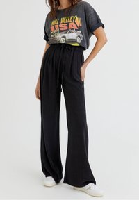 Osoba w czarnych szerokich spodniach z paskiem ściągającym, w vintage'owej koszulce graficznej Hill Valley USA oraz białych sneakersach.