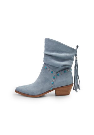 Bottine en daim bleu clair avec tige repliée, petit talon en bois, clous turquoise et argentés, et un détail de pompon à l’arrière.