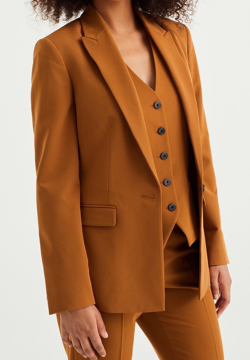 Brun kavaj med skräddarsydd passform, slagkrage, fyra svarta knappar och en enda framficka. Tillverkad av ett slätt tyg med matt finish.