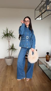 Γυναίκα με τζιν μπουφάν και τζιν παντελόνι με φαρδιά μπατζάκια, κρατώντας μια αφράτη μπεζ τσάντα, στέκεται σε δωμάτιο με φυτό και γυάλινο τραπεζάκι καφέ.