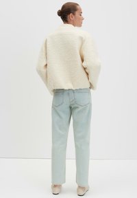 Crème jas met een luchtige textuur en een relaxte pasvorm, voorzien van afhangende schouders. Gecombineerd met lichtblauwe, rechte jeans en beige schoenen.