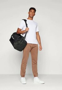 Crni torbu s logotipom, drži osoba u bijeloj majici i bež trenerci, prikazujući jednostavan, sportski dizajn.