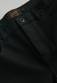 Fekete farmernadrág gombos záródással, textúrált anyaggal és látható varrással. Logófolt a derékrészen, az alap öt zsebes kialakítás.