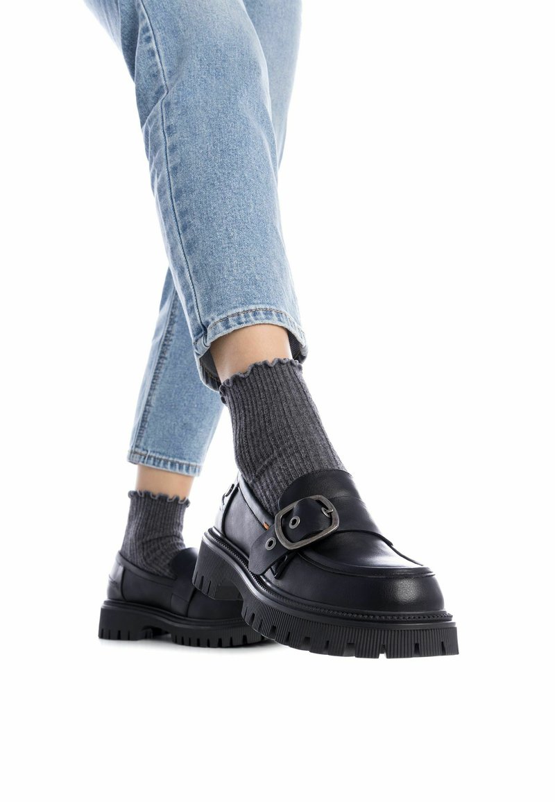 Mocasines de cuero negro con una hebilla, suela de plataforma y fondo de goma texturizado, combinados con calcetines de tobillo acanalados grises y jeans cortos azul claro.
