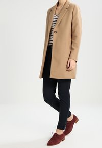 Cappotto di lana color nocciola con un solo bottone, abbinato a una maglietta a righe e pantaloni neri slim-fit. Scarpe in suede bordeaux con lacci completano il look.