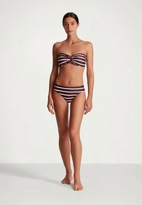 Sieviete stāv basām kājām, tērpusies brūnā un baltā svītrainā bandeau peldkostīmā, uz vienkārša baltā fona.