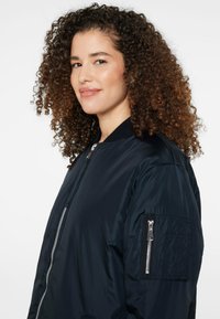 Marineblauer Bomberjacke mit durchgehendem Reißverschluss, geripptem Kragen und Reißverschlusstasche auf dem linken Ärmel. Glatte Textur und schlanke Silhouette.