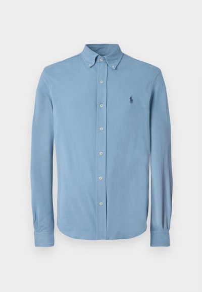Svetlo modra srajca z gumbi, izdelana iz gladkega materiala, z klasičnim ovratnikom, dolgimi rokavi in majhnim temnim grbom na prsih.