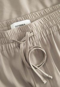 Kreminės spalvos džinsai su elastine juosta, raišteliu ir etiketė su užrašu "GABBA." Lygus paviršius, paprastas dizainas, nematyti raštų.