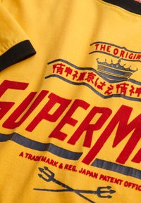 Yellow cotton t-shirt featuring bold red and blue graphics, including "SUPERM" and a crown design, with black trim on the collar.
