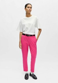 T-shirt blanc surdimensionné, pantalon rose vif sur mesure avec poches, ceinture noire, chaussettes grises et mocassins noirs. Textures de tissu lisses.