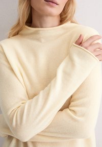 Jasnożółty, miękki sweter z wysokim dekoltem i długimi rękawami, o luźnym kroju i lekko teksturowanym wykończeniu.