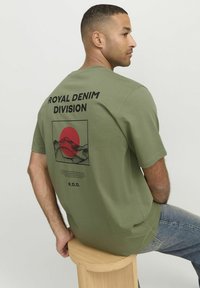 Camiseta de manga corta verde oliva con un estampado gráfico negro en la espalda que muestra montañas y un sol rojo, etiquetada "Royal Denim Division."