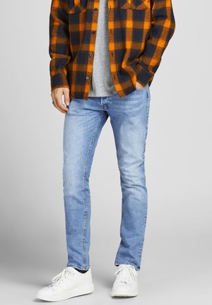 Homme portant une chemise à carreaux orange et noir, un jean bleu clair, des baskets blanches, se tenant devant un fond gris.