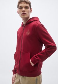 Červená mikina na zips z mäkkého materiálu s predným klokankovým vreckom a rebrovanými manžetami. Na hrudi má zlaté vyšívané logo.