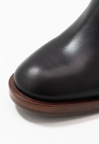 Bota negra de cuero con textura lisa, punta redonda y suela de madera marrón, sin patrones ni detalles de herrajes notables.