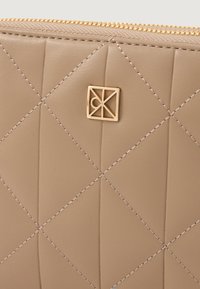 Beige læderpose med diamantquiltet mønster, gyldent hardware og en geometrisk logo detalje. Lynlåslukning på toppen.
