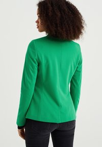 Grön kavaj i slät tyg med en figursydd design och långa ärmar. Bakdetaljen inkluderar en mittsöm och subtila ventiler.