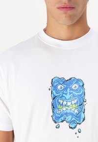 Camiseta blanca con un diseño gráfico azul de una cara rompiendo la tela, con detalles intrincados y textura visible.
