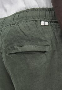 Olivgrüne Leinen-Shorts mit elastischem Bund, einer kleinen Gesäßtasche und subtiler Struktur. Ein weißes Etikett ist in der Nähe des Bundes angebracht.