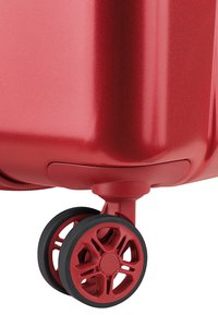 Valigia rigida rossa con superficie liscia e lucida, dotata di una ruota nera multi-direzionale con design a raggi geometrici.
