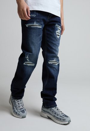 Person iført mørkeblå revnede jeans og grå atletiksko, stående mod en ensfarvet lys grå baggrund.