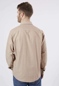 Chemise longue beige clair à manches longues avec col, dotée de coudières, de coutures contrastées et d'un ourlet arrondi. Vue de dos présentée.