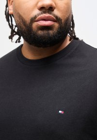 Κοντινό πλάνο άνδρα με γένια που φοράει μαύρο πουκάμισο με ένα μικρό κόκκινο, λευκό και μπλε ορθογώνιο λογότυπο στο στήθος.