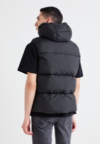 Čierny puff vest s vysokým golierom a veľkou kapucňou, s prešívanými časťami a hladkou, lesklou textúrou. Nosené cez čierne tričko.