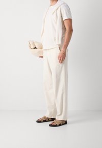 Beige kortärmad skjorta, lösa cremefärgade byxor, tanfärgade sandaler med korsade remmar, håller en ihoprullad beige tröja. Enfärgad bakgrund.