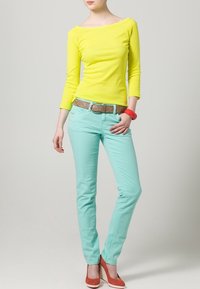 Jasně žlutý top bez ramínek, mentolově zelené slim-fit kalhoty, hnědý opasek s průvleky a korálové podpatky. Hladké textury a živé barvy.