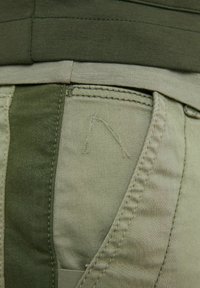Pantaloni verde oliva caratterizzati da diverse trame nel tessuto. Dettagli di cucitura notevoli in un colore più chiaro, con un patch logo ricamato sopra la tasca.