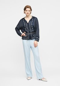 Γυναίκα που φοράει πουκάμισο με μπλε-θαλασσί σχέδια και ζώνη με αλυσίδες, καθώς και ανοιχτό μπλε παντελόνι με φαρδιά πόδια, στέκεται με το ένα χέρι στη μέση μπροστά από λευκό φόντο.