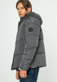 Šedá péřovka s kapucí, prošívaný design, boční zip a okrouhlá nášivka na levém rukávu. Hladký, lesklý materiál s uvolněným střihem.