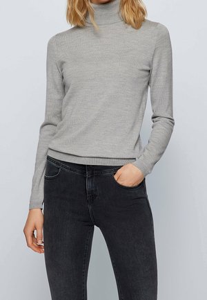 Žena nosící přiléhavý světle šedý rolák a černé džíny s vysokým pasem, s jednou rukou v kapse, na jednoduchém pozadí.