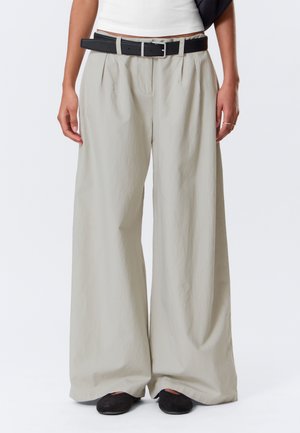 Personne portant un pantalon large beige ample avec des plis, une ceinture noire, un haut court blanc et des chaussures noires sans lacets, debout devant un fond uni.