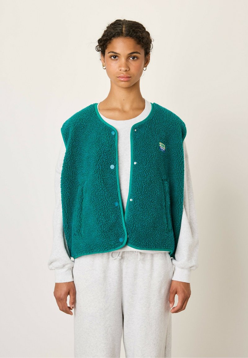 Teal fleece vest met een gestructureerd oppervlak, drukknoopjes, contrasterende afwerking en een klein logo op de linkerkant van de borst, gedragen over een grijze sweatshirt.