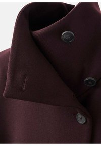 Bordo vuna kaput s visokim ovratnikom, dva crna gumba i malim detaljem bočne džepne. Glatka tekstura s strukturiranim dizajnom.