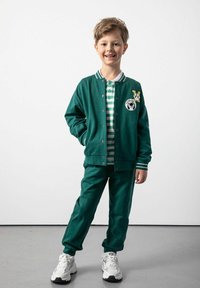 Zielony dres z tkaniny, z paskowanym polo pod spodem. Obejmuje sportową kurtkę z naszywką z logo i białymi paskami. Białe sneakersy.