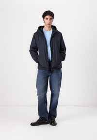Marineblauwe hoodie-jack met een gladde textuur, gedragen over een lichtblauw shirt, gecombineerd met lichtblauwe jeans en zwarte schoenen. Minimalistisch ontwerp.