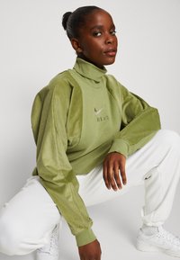 Grön Nike-pullover med hög krage, med ribbad textur på ärmar och bröst. Modellen bär vita byxor och poserar i en hukande ställning.