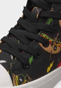 Κοντινή λήψη ενός μαύρου αθλητικού παπουτσιού με χοντρές μαύρες κορδόνια και ζωντανά καρτούν μοτίβα σε πράσινο, κόκκινο, κίτρινο και μπεζ στο ύφασμα στην επάνω πλευρά.